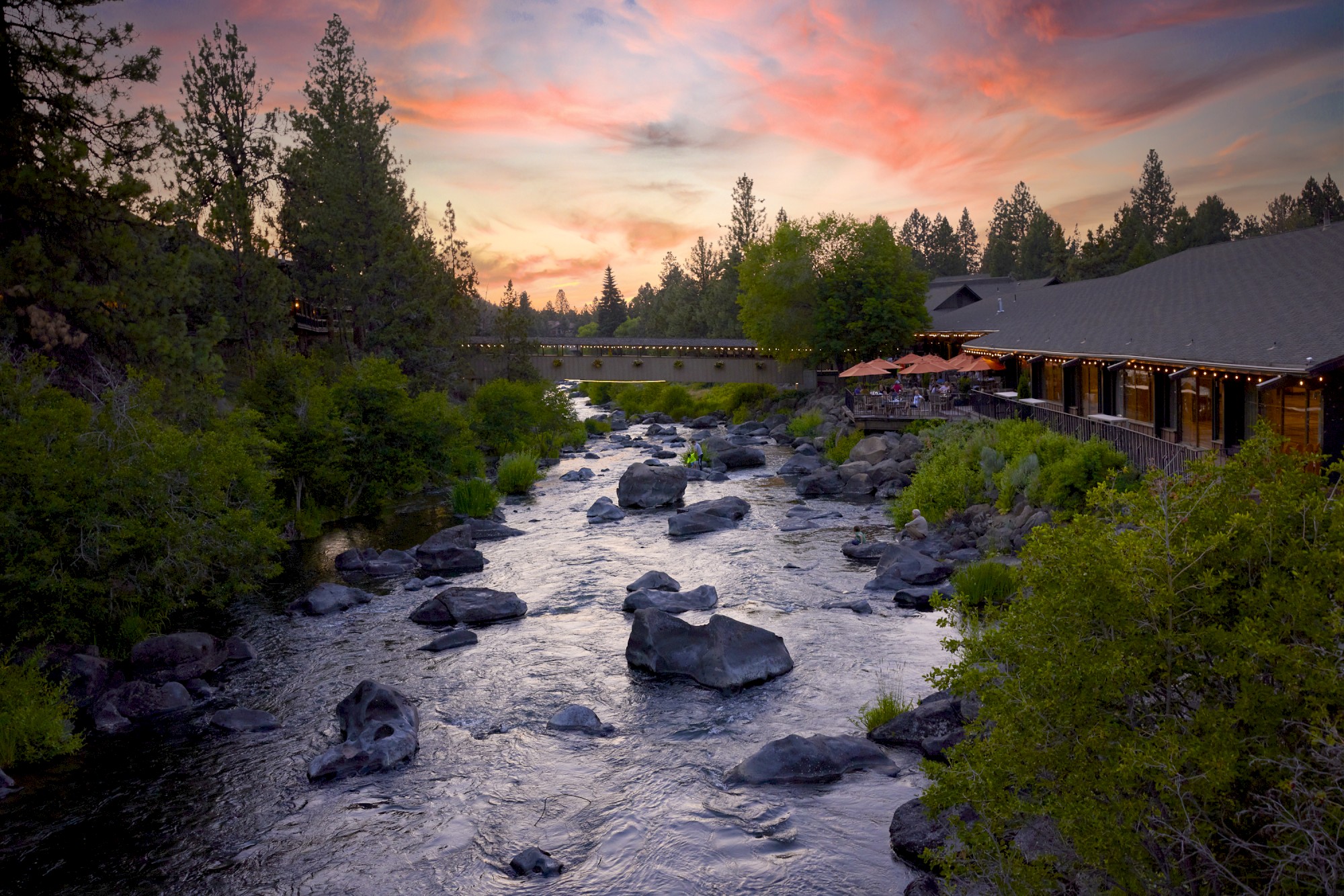 Rippling river with rocks, trees, sunset sky, and Riverhouse in Bend, Oregon on the right side.