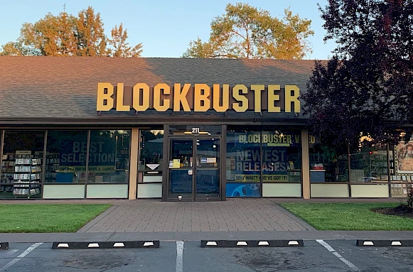 The image shows The Last Blockbuster, Bend Oregon with a large yellow sign and promotional posters in the windows. Photo Credit: Wikipedia